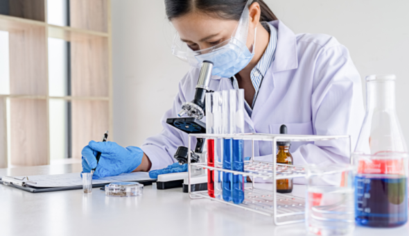 Person in a lab coat looking into a microscope doing an experiment in a laboratory. There's a row of test tubes on the bench. The person is writing on a clipboard.