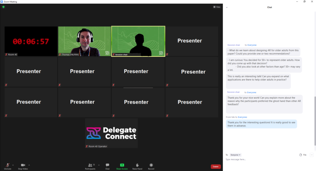 A screenshot of the presenter's experience. A Zoom meeting with 13 participants, shown in gallery view. I have my video on, and the chair of the session's video is displayed but with the presenter blacked out for anonymity. The top left participant is a count down, controlled by the service providing the technical support, Delegate Connect, whose logo is visible as another participant. All other presenters (9 in total) are anonymous authors with their cameras off. The chat is open and a sample of questions that I was asked by the session chair are visible. The questions are: 1. What do we learn about designing AR for older adults from this paper? Could you provide one or two recommendations?  2. I am curious: You decided for 50+ to represent older adults. How did you come up with that decision? 3. Did you also look at other factors other than age? 50+ may vary a lot.  4. This is really an interesting talk! Can you expand on what applications are there to help older adults in practice? 5. Thank you for your nice work! Can you explain more about the reason why the participants preferred the ghosthand than other AR feedback?  A final comment from me reads: Thank you for the interesting questions! It is really good to see them in advance. 