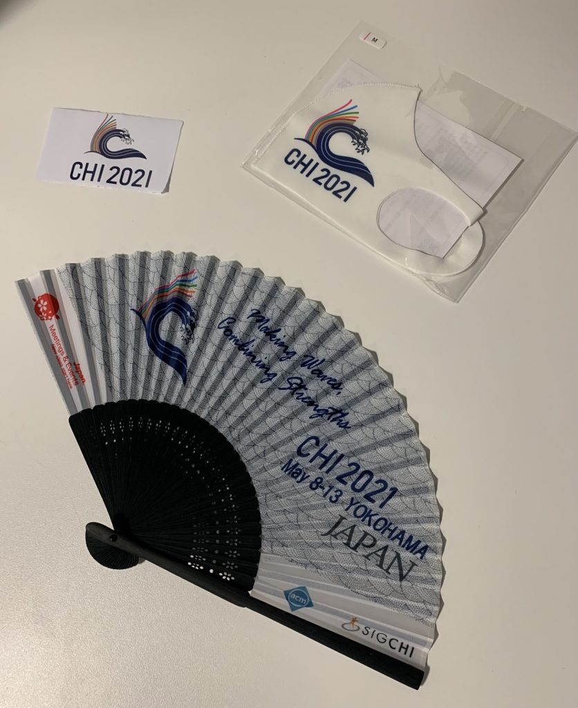 A picture of some items with CHI branding on them, including a piece of paper with the CHI logo, a CHI face mask, and a CHI paper fan. 