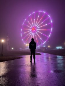 A person stands before a colourful and purple fair wheel