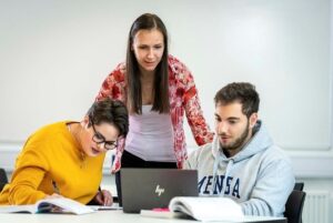 Tutor standing, discussing with two seated students, around a laptop