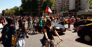 A large group of demonstrators, some masked, walk down a road infront of tower blocks. One has a Chilean flag.