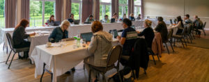 A group of students and teachers sit around a U shaped table writing.