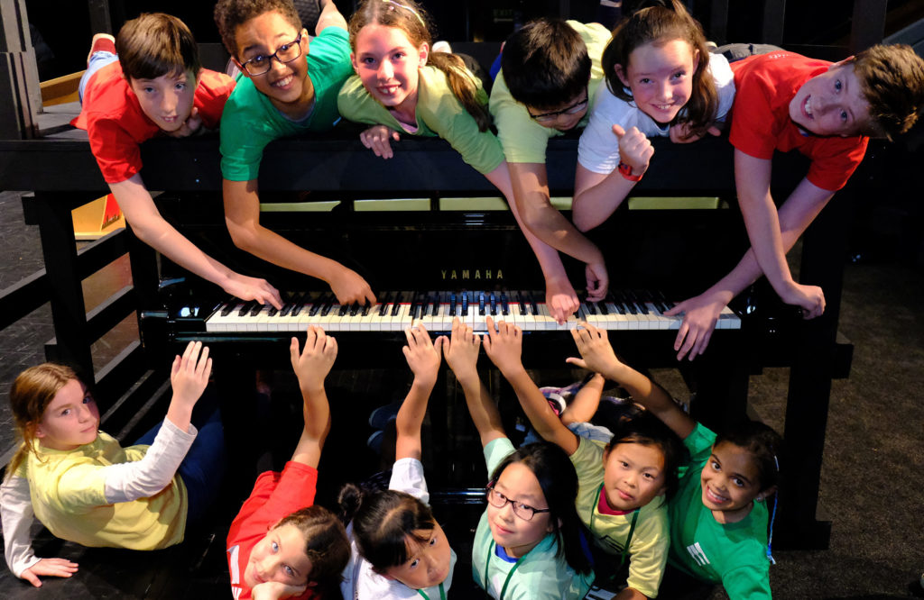 Some children are surrounding a piano happily and posing in front of the camera.