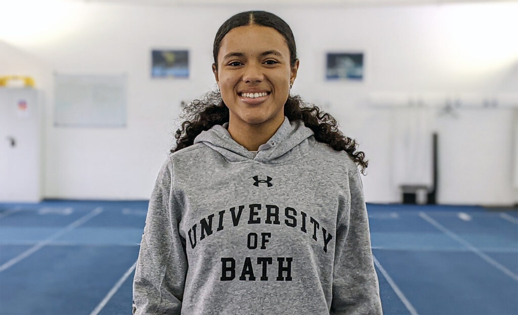 Black girl with curly hair in a ponytail wearing a grey University of Bath hoodie and smiling.