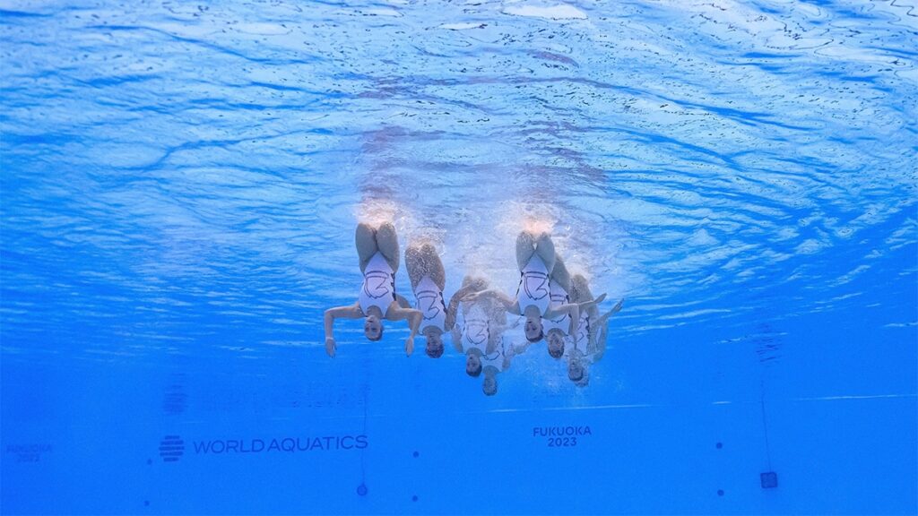 Eight women under the surface of the water upside down with their legs sticking above the pool.