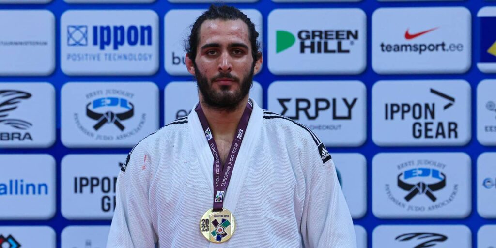 Man with dark hair and a full beard in a white judo kit wearing a gold medal.