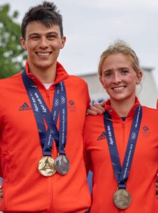 A brunette white man and a blonde white woman smiling, wearing bright orange athleisure wear and medals around their necks.