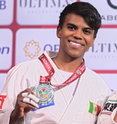 Brown woman with short black hair wearing a judo uniform with the Mexican flag on the chest, holding a silver medal and grinning.