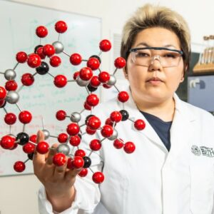 A Kyrgyz woman with short blonde hair and safety glasses holds a model of a molecule aloft. 