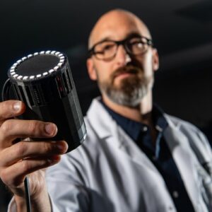 A bald, bearded man wearing a labcoat and glasses holds a black machine proudly in his hand.