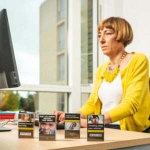 A white woman with short blonde hair and a yellow cardigan sits at a desk looking at a computer. There are unopened packets of cigarettes on her desk. 