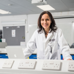 A brunette woman in a lab coat stands in front of a computer and smiles.