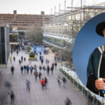 An old man in a ceremonial graduation cap and gown holding a degree and smiling against of backdrop of the University of Bath Parade street.