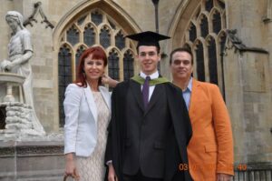 Martin, wearing graduation cap and gown on his graduation day. He's between his mother (left) who has red hair and is wearing a cream dress, his father (right) is wearing an orange suit.