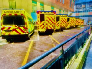 Seven ambulances are lined up and closely parked outside a city centre hospital. The ambulances are bright yellow with red stripes. The brightness and contrast convey a halo effect on the ambulances.