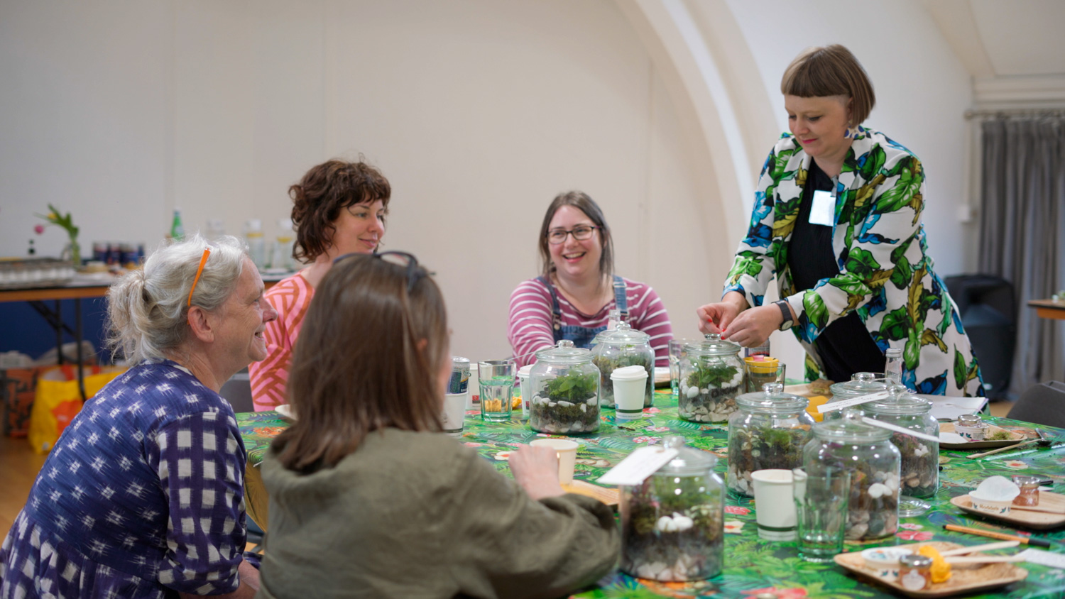 Five people sit around a table creating terrariums together. They appear deep in conversation.