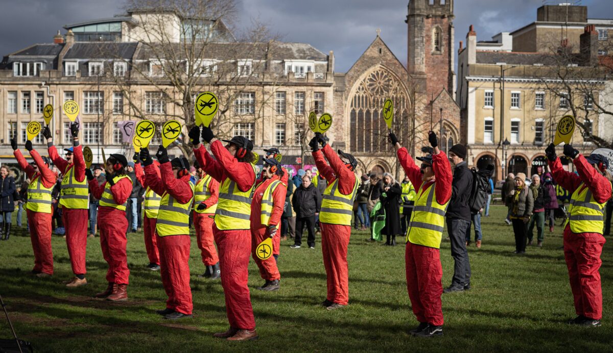 This image is of a group of 12 protesters dressed as air traffic controllers in high-vis vests holding up paddles with a vector of an airplane crossed out, stand on College Green, campaigning against the expansion of Bristol Airport. Several members of the public stand behind them looking on to the protest.
