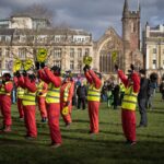 This image is of a group of 12 protesters dressed as air traffic controllers in high-vis vests holding up paddles with a vector of an airplane crossed out, stand on College Green, campaigning against the expansion of Bristol Airport. Several members of the public stand behind them looking on to the protest.