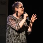 A woman holding a microphone gesturing to the audience in front of a back background. There is a banner for the 'Minerva Lecture Series' behind her.