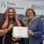A woman with long pink hair holding a certificate with a woman in a blue top. Both are smiling at the camera.