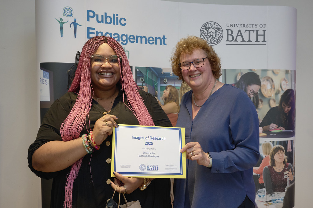 A woman with long pink hair holding a certificate with a woman in a blue top. Both are smiling at the camera.