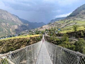 The images show a metal suspension footbridge stretching across a valley. On the other side is a rural, hilly village in the Mid-Western region of Nepal. There are a few buildings and terraced fields with lots of trees. On the bridge is a woman dressed in red, she is holding on to the left side of the bridge and looking out into the distance. The sky is sunny but directly across the bridge there are dark clouds approaching. 