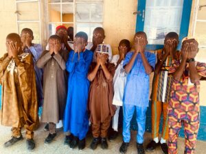 A group of Nigerian secondary school students stand together in their cultural attires with their faces partially or fully covered by their hands. Their body language conveys a sense of silence and concealment. The background is a school setting. The image symbolises the suppression of students’ linguistic and cultural identities due to school policies that ban Indigenous languages and prioritise English. It visually represents the quiet resistance of students in a system that punishes them for speaking their mother tongues. 