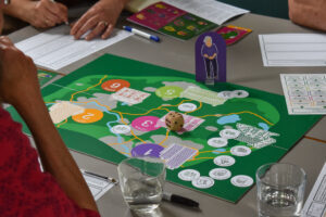 We see three people sitting around a table; just their arms are visible. They are playing a board game. There is a board with a stylised map showing city, town, and other locations, a stand-up image of an older woman, dice, and other game elements that are unclear. 