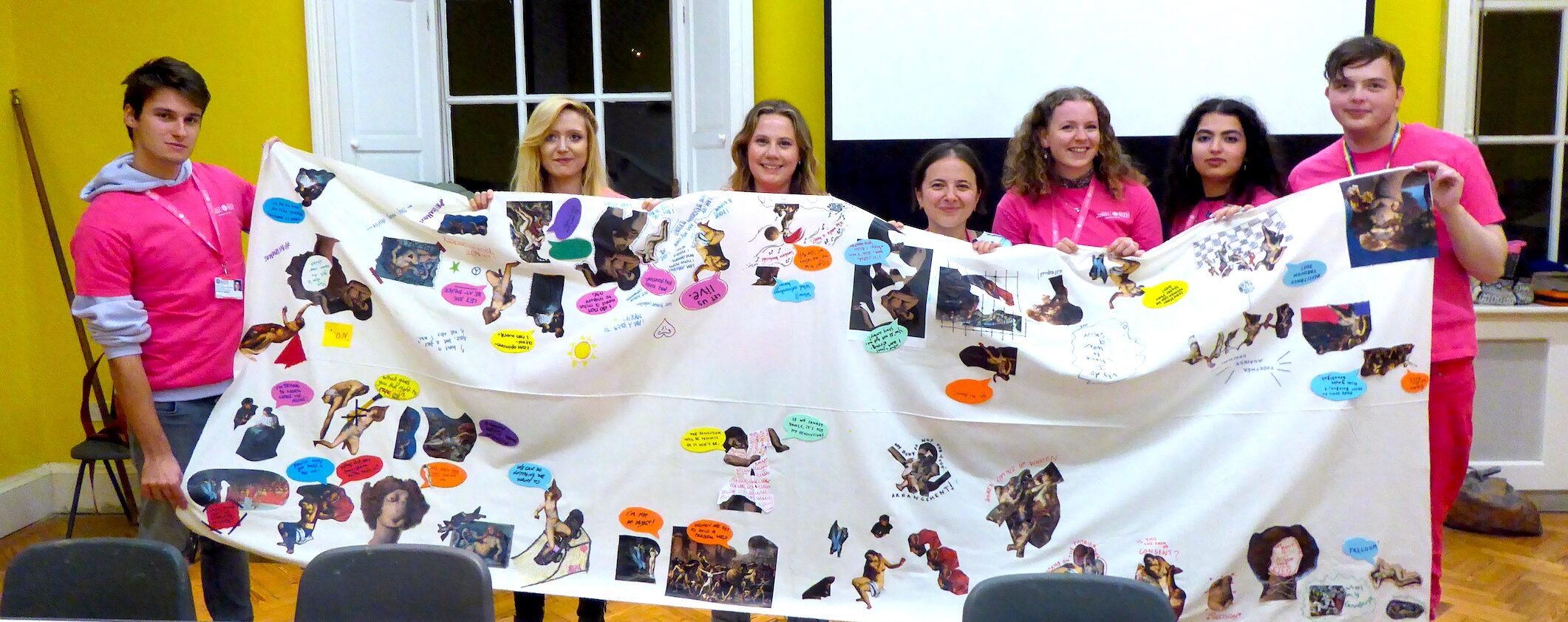 Seven people in pink festival t-shirts hold up a large white banner full of images and text about women in art and museums