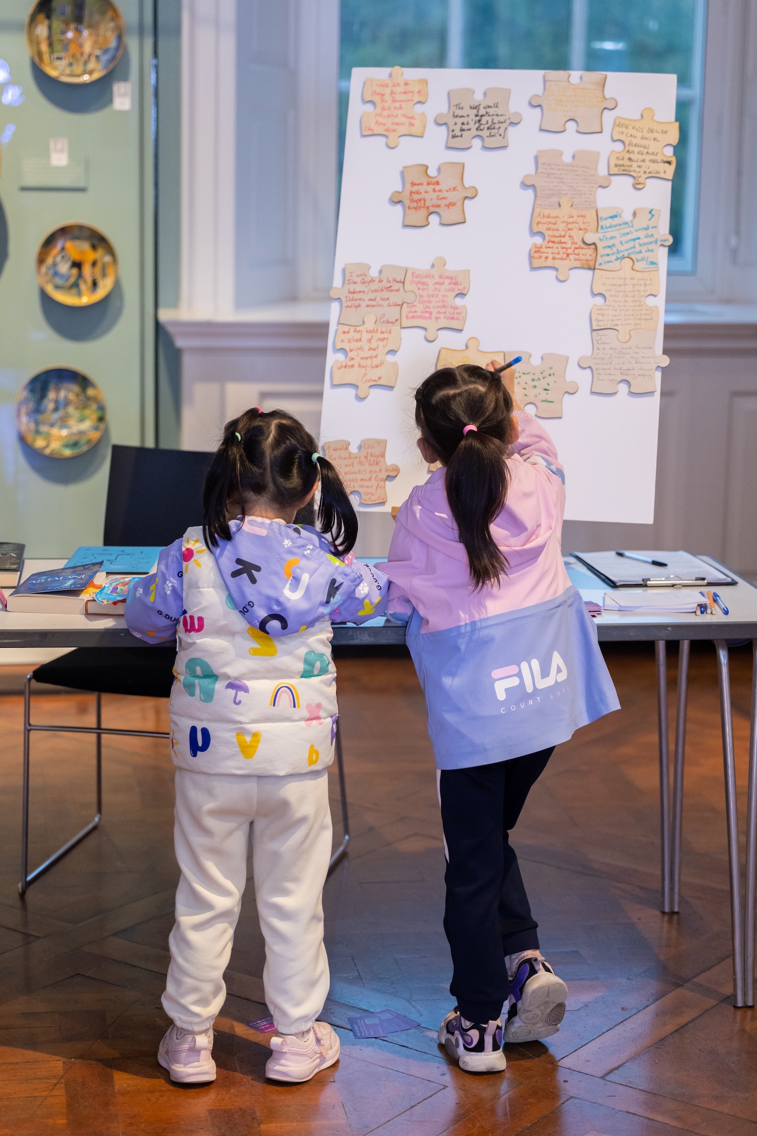Two young people lean on a table in a museum, writing their thoughts on large wooden jigsaw pieces to add to a display