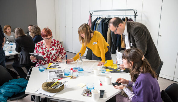 A group of people standing around a table. The table has post it notes and pens on it.
