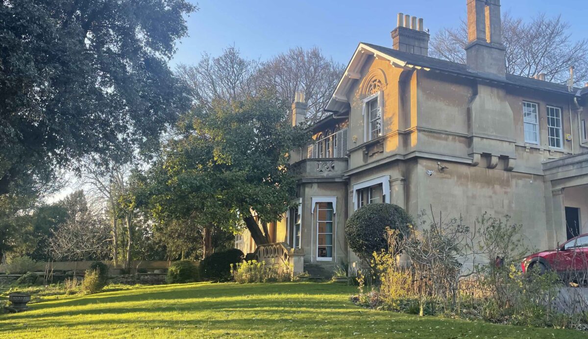 Fairfield House, a pale two story house with large chimneys, lit with golden sunlight, situated in an open garden with green lawn and large mature trees.
