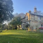 Fairfield House, a pale two story house with large chimneys, lit with golden sunlight, situated in an open garden with green lawn and large mature trees.