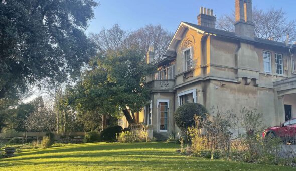 Fairfield House, a pale two story house with large chimneys, lit with golden sunlight, situated in an open garden with green lawn and large mature trees.