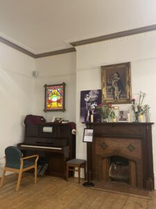 Room with white walls are wooden floor. An upright mechanical piano is situated in the corner, next to an open fire place. 