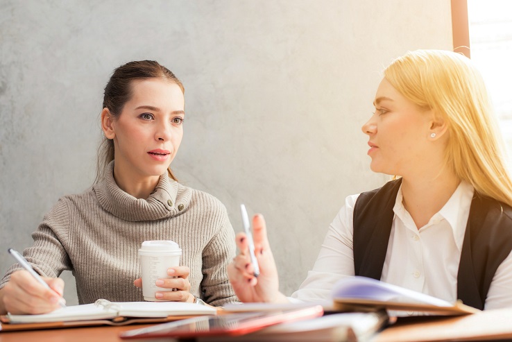 Two women sitting side by side, both are holding a pen and have a notebook open. One is also holding a coffee.