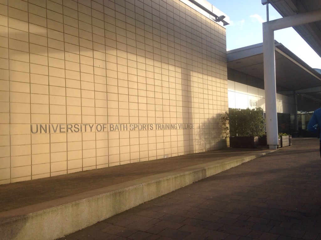 Brick wall with the words UNIVERSITY OF BATH SPORTS TRAINING VILLAGE on it. The entrance to the STV is to the right.