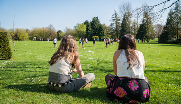 Two female students sat cross-legged on some grass watching a group of students playing football