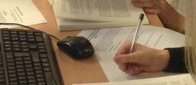 Image of someone's hand writing with a pen with a keyboard and mouse on the right-hand side