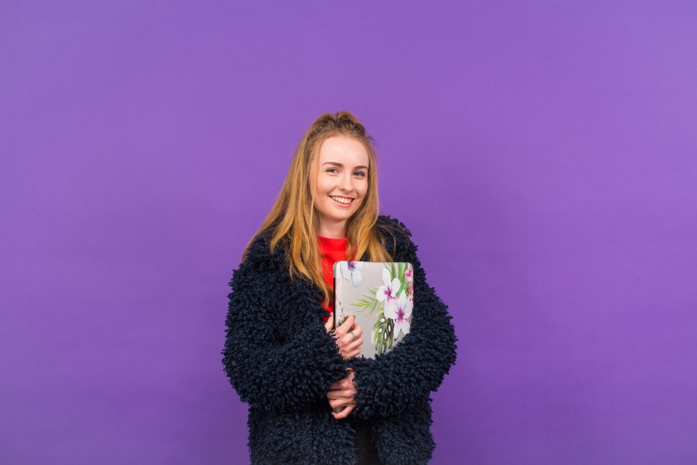 Ellie stood against a purple background holding a flowered laptop