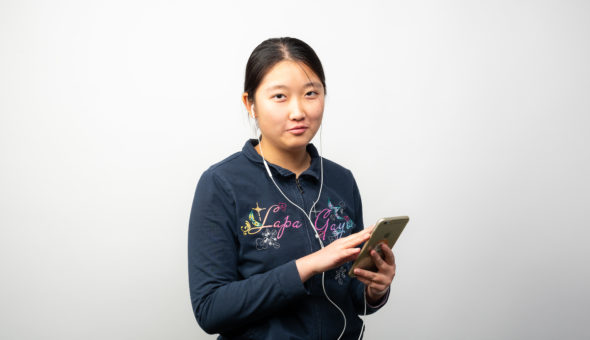A photo of Lily wearing some white headphones attached to a gold iPhone. She is wearing a navy blue zip up sweatshirt with colourful writing on the front.