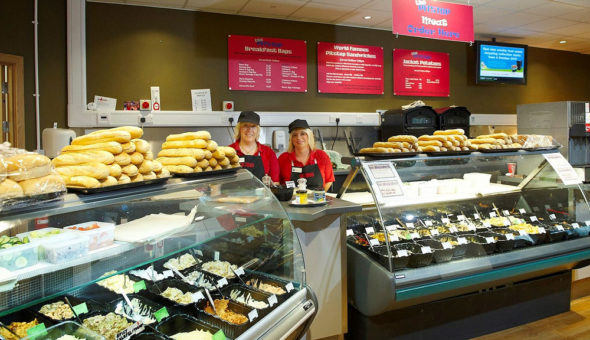 Two ladies wearing red and black head covering stood behind a deli counter. There are losts of sandwiches and baguettes on display in the glass counter.