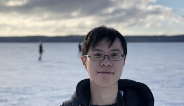 Photo of Duncan who is East Asian. He is wearing black metal glasses and black clothing and is surrounded by snow on the the ground in the background. There are two people who are out of focus in the background.