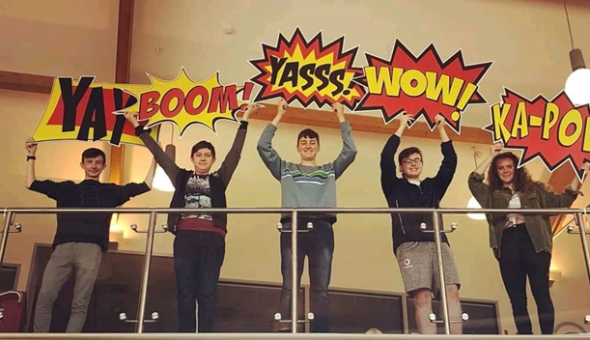 5 males students stood in front of a clear glass railing. Each student is holding a sign above their head. The signs say Yay, Boom,Yass, Wow and and Yass, Wow and Ka-Po(w). The latter 'w' is not showing. The signs are in 'cartoon-style' writing with jagged edges in bright red, yellow and black.