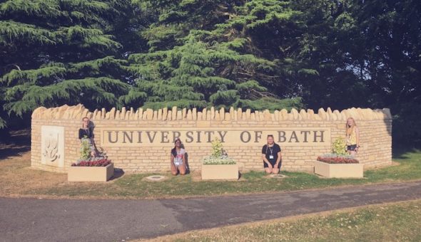 Four female students outside the main Unversity of Bath stone wall entrance. Two are stood up and two are kneeling down and they are all in front or t oteh side of three stone raised beds. There are green trees in the backgound and a grey stone pathway runing the length of the photo which is in front of them.