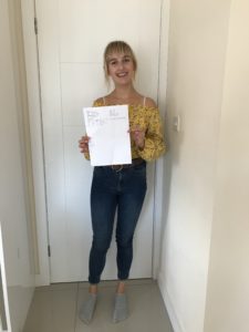 Full- length photo of me holding my A Level results paper. I'm stood in a corridor with my back against a white door which is closed. 