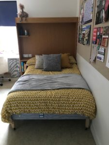 Portrait photo of a bed with yellow duvet and grey and yellow cushions and grey throw. There is a notice board with lots of notices pinned to it on the righthand side.