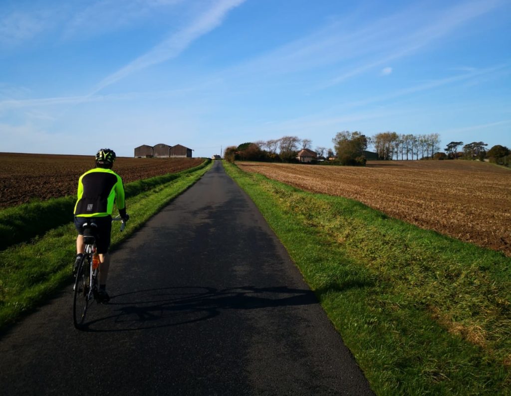 I am cycling on an empty road. There is a green grass bank and brown fields either side of the road which has no road marking. There are trees, three farm buidlings and a farm house in the distant top of the photo. The sky is blue with some long straight clouds.