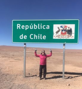 Rachel standing standing under a green and white sign at the Chilean border which says Republica de Chile. There is also a logo on the sign.